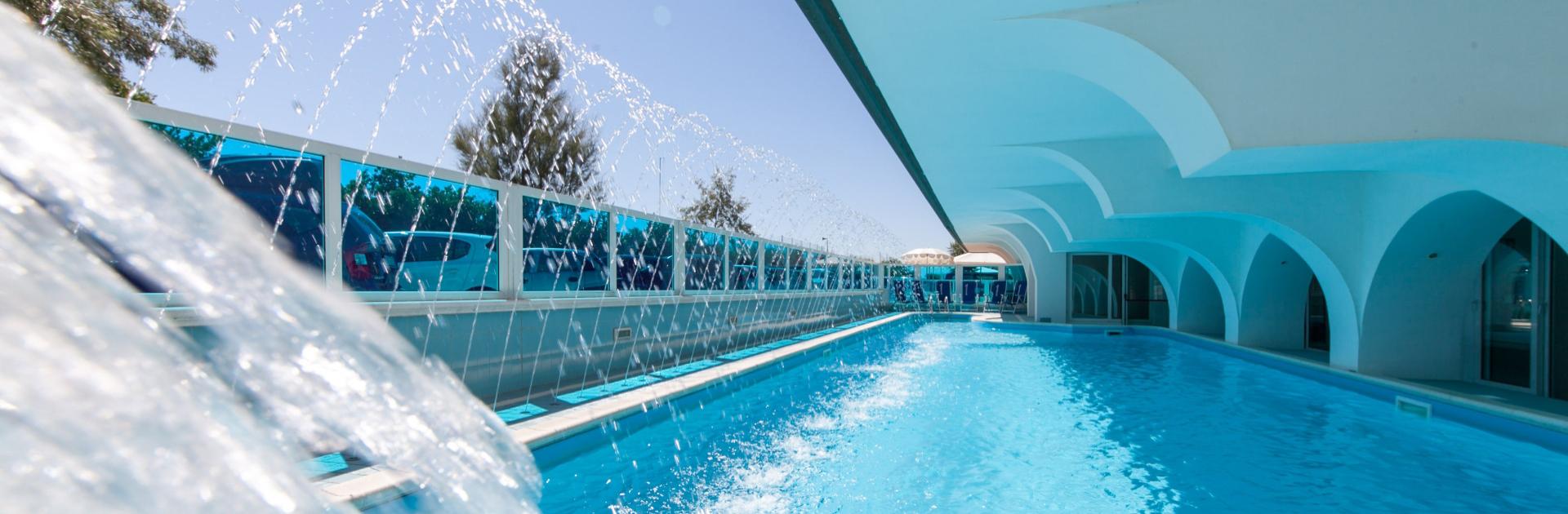 Piscina coperta con archi bianchi e giochi d'acqua, ambiente luminoso e moderno.